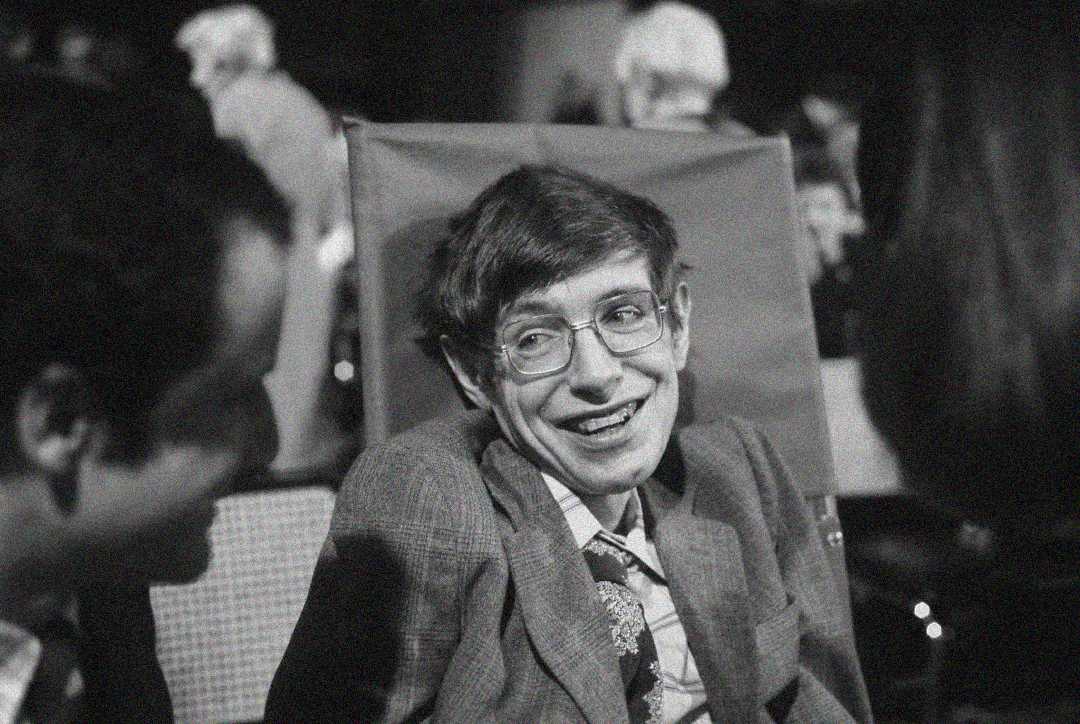 A man in a wheelchair wearing glasses and a suit smiles while sitting among others in a crowded indoor setting. The image is black and white.