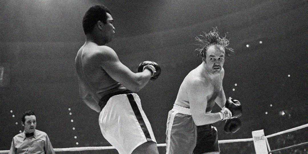 Two male boxers are fighting in a ring. One boxer is landing a punch, making the other boxer’s head snap back and hair fly. A referee stands in the background, watching the match.