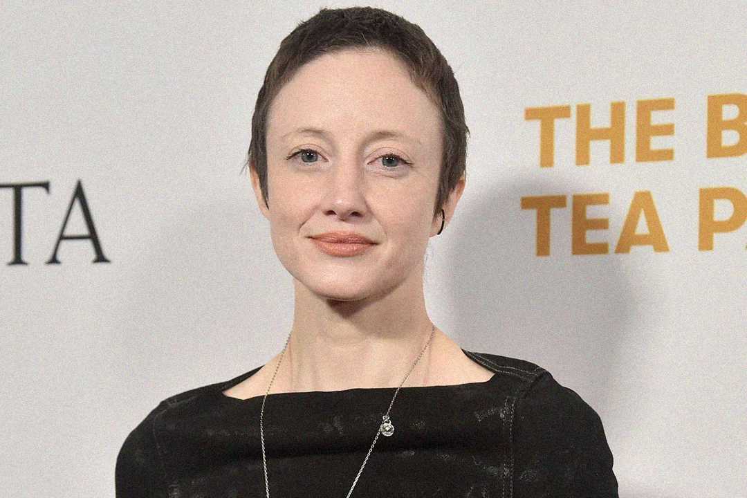 A woman with short brown hair, wearing a black top and a silver necklace, stands in front of a backdrop with partial text that reads "THE B... TEA P...".