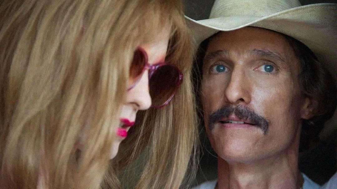 A person in pink sunglasses and long hair is in the foreground, slightly out of focus, while a man in a white cowboy hat and mustache looks on intently in the background.