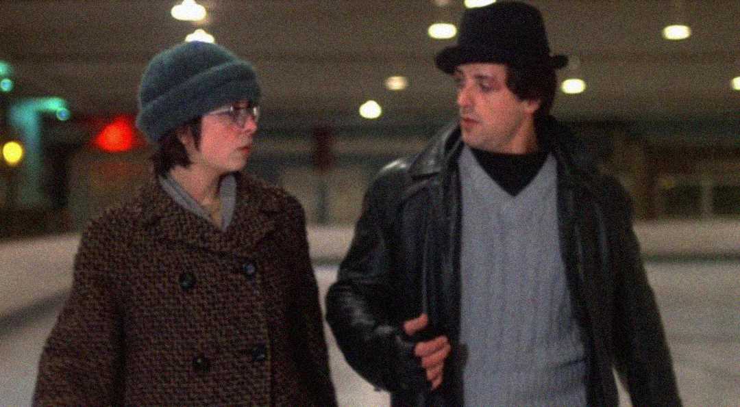 Two people walk side by side in winter clothing, including hats and coats, in what appears to be an indoor ice rink. They are engaged in conversation and looking at each other. The background is softly lit and out of focus.
