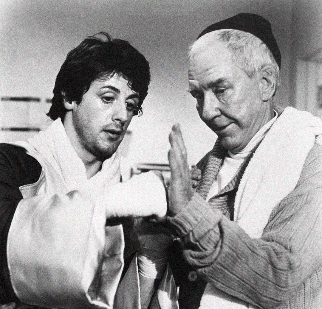 A young man with boxing gloves and wrapped hands receives guidance from an older man. Both are focused, with the older man gesturing as if giving advice. The photo is in black and white.