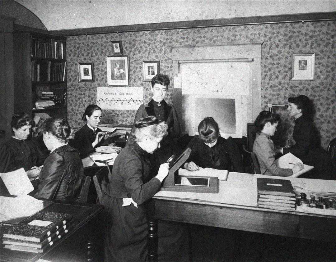 Black and white photo of eight women working at desks with papers and books in a Victorian-era office. Some are reading or writing, while one stands in the center observing. Bookcases and framed pictures decorate the background.