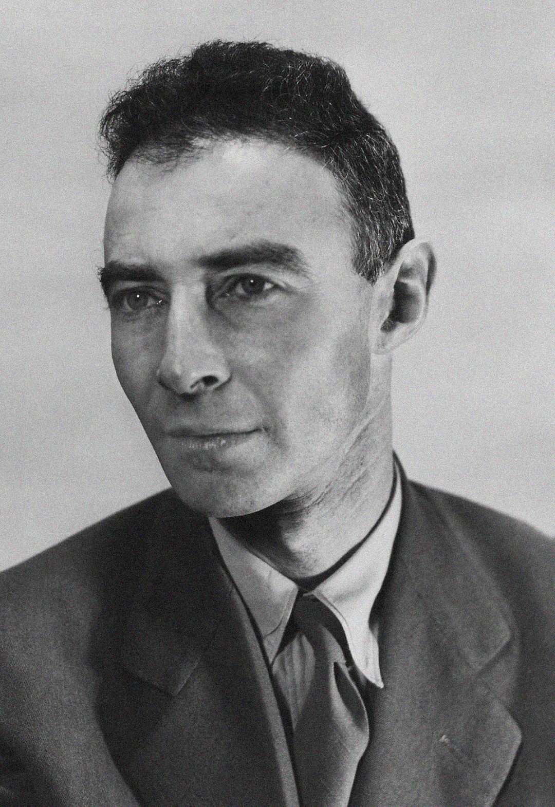 Black and white portrait of a man with short dark hair, wearing a suit jacket, dress shirt, and tie, looking slightly to the side with a neutral expression.