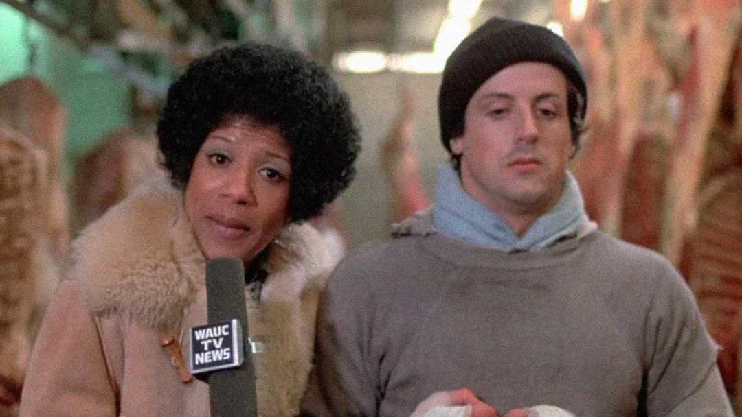 A woman holding a "WAUC TV NEWS" microphone interviews a man in a hoodie and beanie. They stand close together indoors, with blurred shapes and hanging objects in the background.
