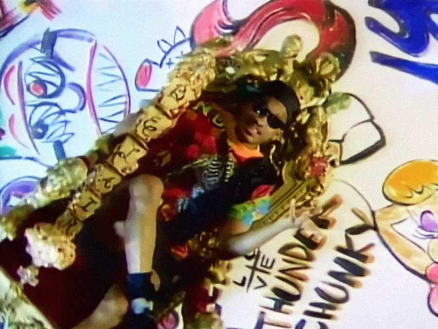 A young man wearing sunglasses and colorful clothes reclines on an ornate golden throne. Graffiti-style artwork and bold text cover the wall behind him.