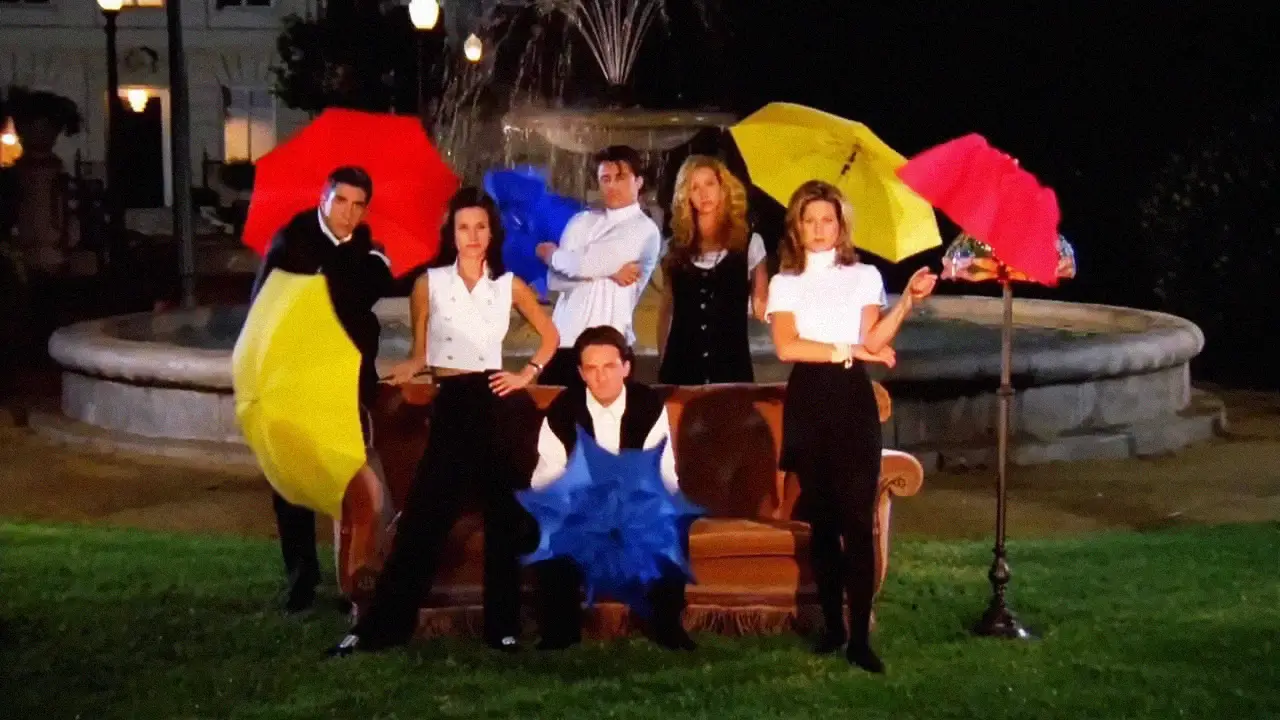 Six people pose in front of a fountain at night, holding colorful red, yellow, and blue umbrellas, standing and sitting around a brown couch on a grassy lawn.