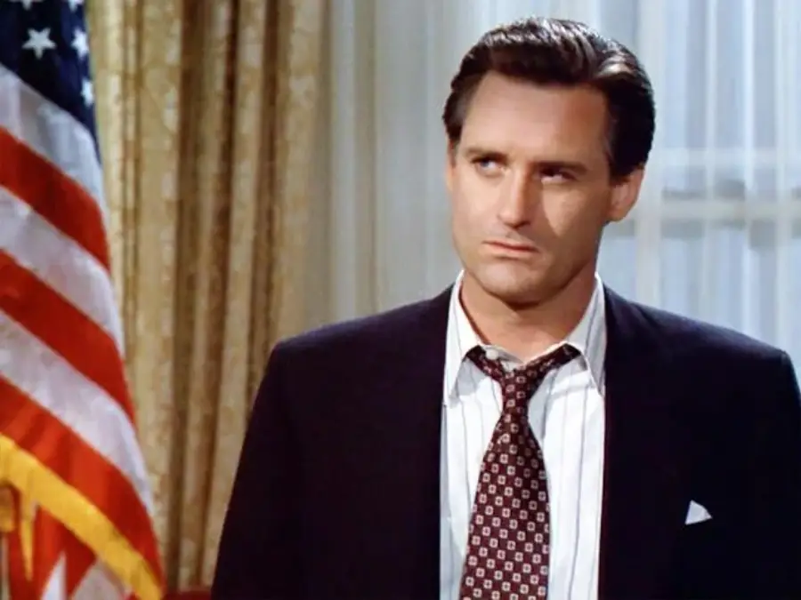 A man in a suit and tie stands indoors beside a large American flag. He has short, neat hair and a serious expression, with curtains and a window in the background.