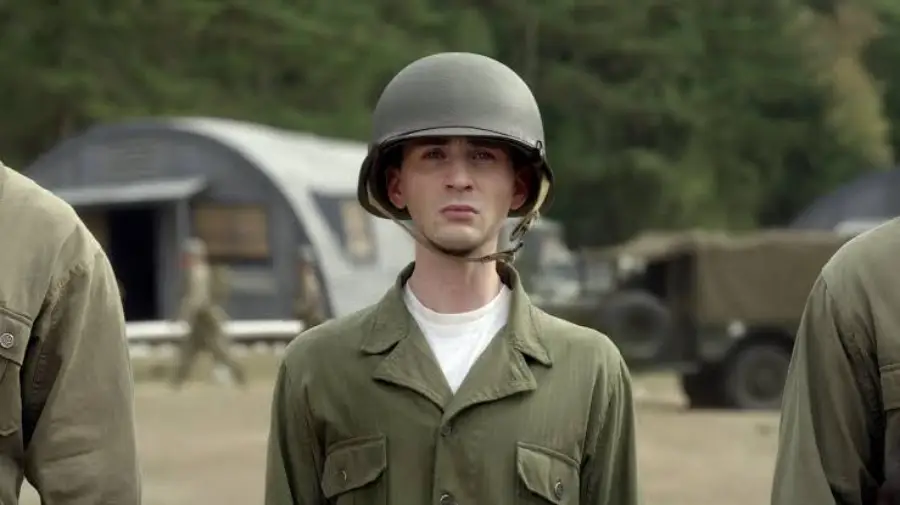A young man in an oversized military uniform and helmet stands outdoors at a training camp, with barracks and vehicles visible in the background. He appears serious and focused.