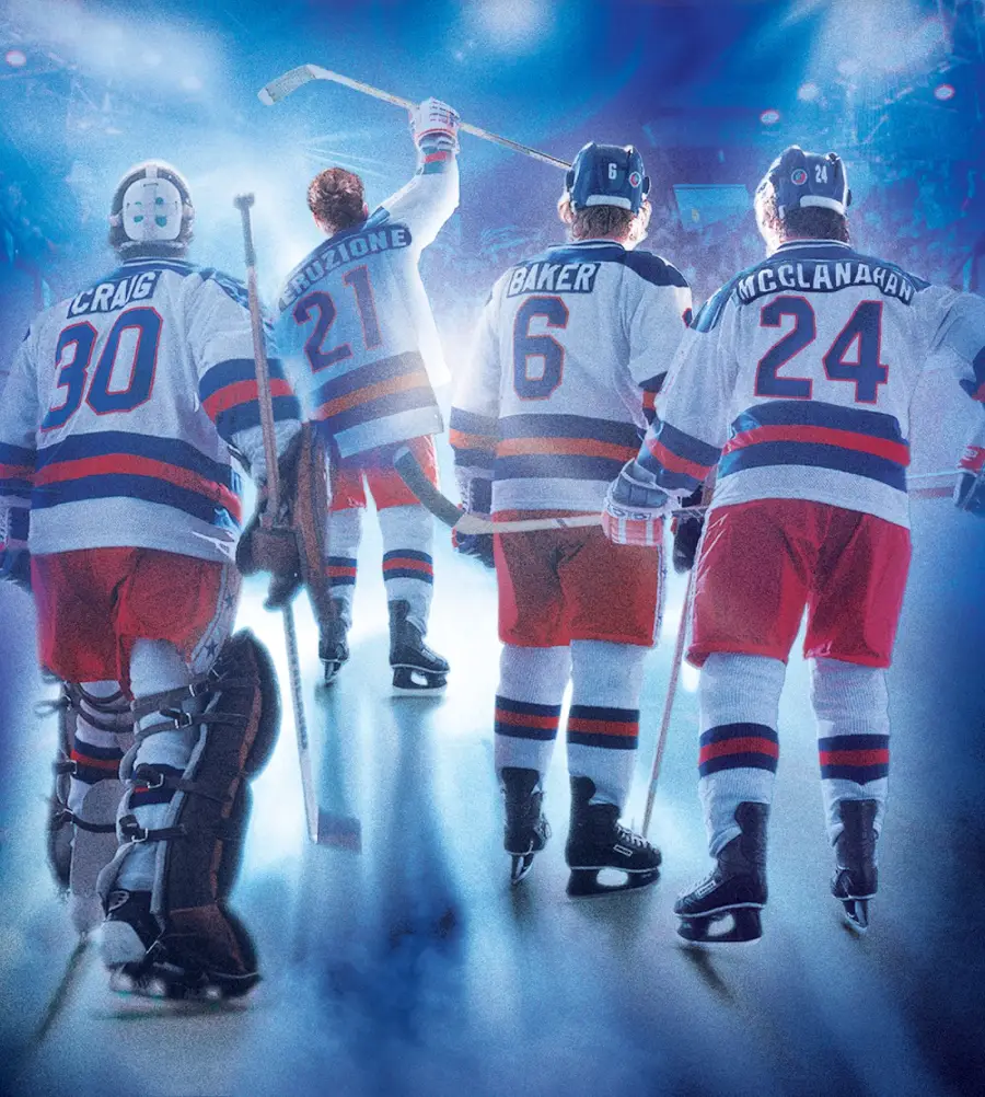 Four ice hockey players in red, white, and blue uniforms walk on the rink toward a cheering crowd, seen from behind. One player raises a hockey stick in celebration under bright arena lights.
