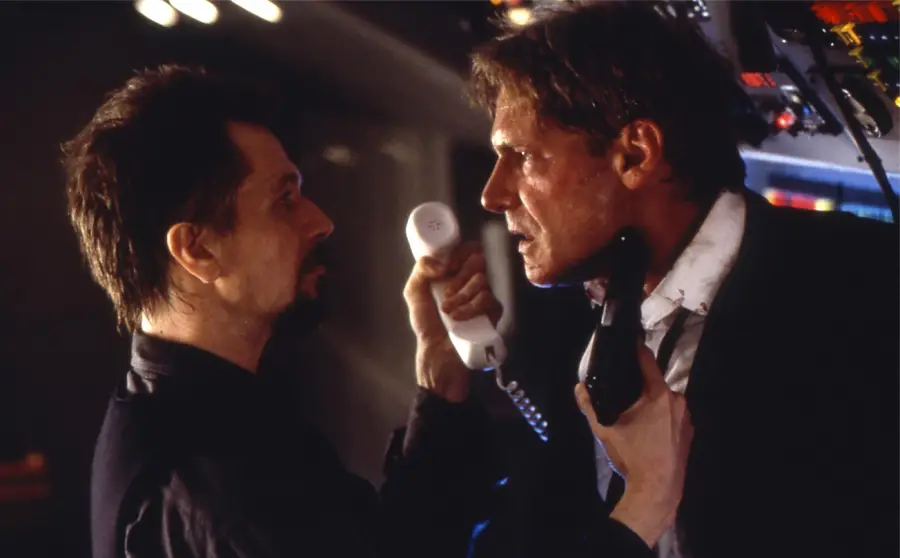 Two men stand face to face in a tense confrontation; one grips the other’s collar and holds a white phone, while the other holds a black phone. They appear intense and sweaty, standing in a dimly lit control room.
