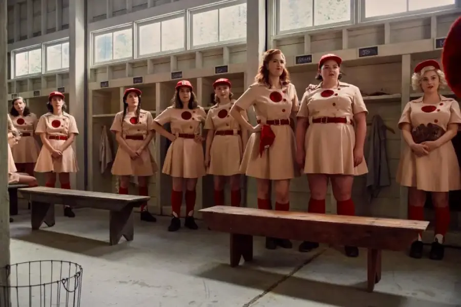 A group of women in vintage baseball uniforms stand in a row inside a locker room, wearing beige dresses with red belts, red socks, and red caps. Wooden benches and cubbies are visible in the room.