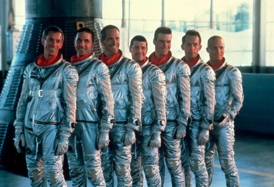 Seven astronauts in shiny silver spacesuits and red neck scarves stand in a line, smiling, inside a hangar with a space capsule in the background and large windows letting in natural light.