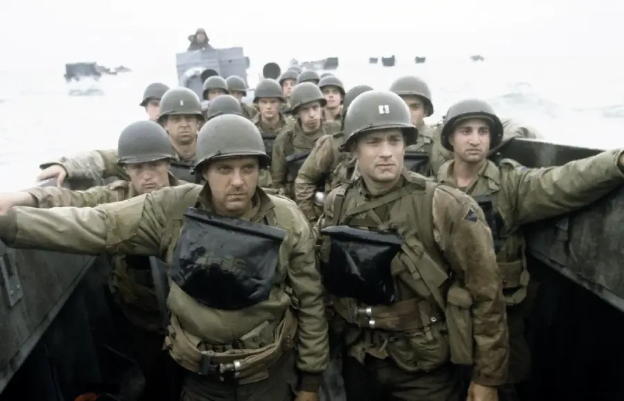 A group of American soldiers in helmets and gear stand closely together in a landing craft, heading toward shore during World War II. The scene is tense and somber, with boats and water visible in the background.