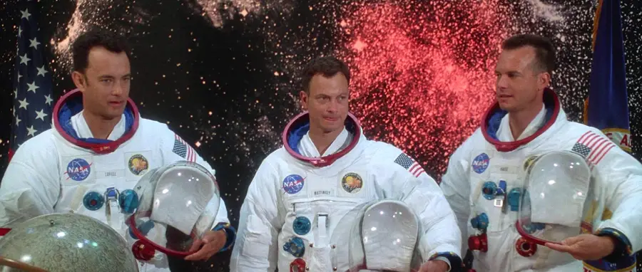 Three astronauts in white NASA spacesuits with helmet visors stand in front of a backdrop showing space and red clouds, holding their helmets and looking in different directions. An American flag is visible to the left.