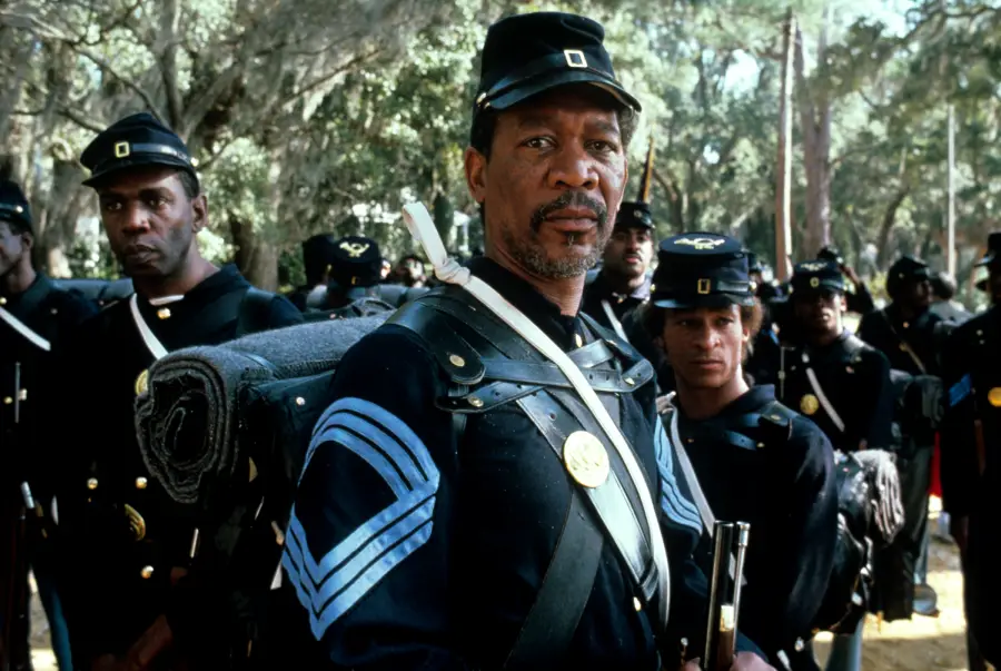 A group of Black Union soldiers in Civil War uniforms stand together in a wooded area; the central figure looks directly at the camera, wearing a hat, carrying gear, and holding a rifle.