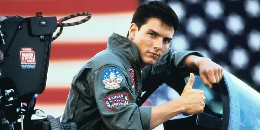 A man in a military flight suit gives a thumbs-up while sitting in a jet cockpit, with a large American flag visible in the background.
