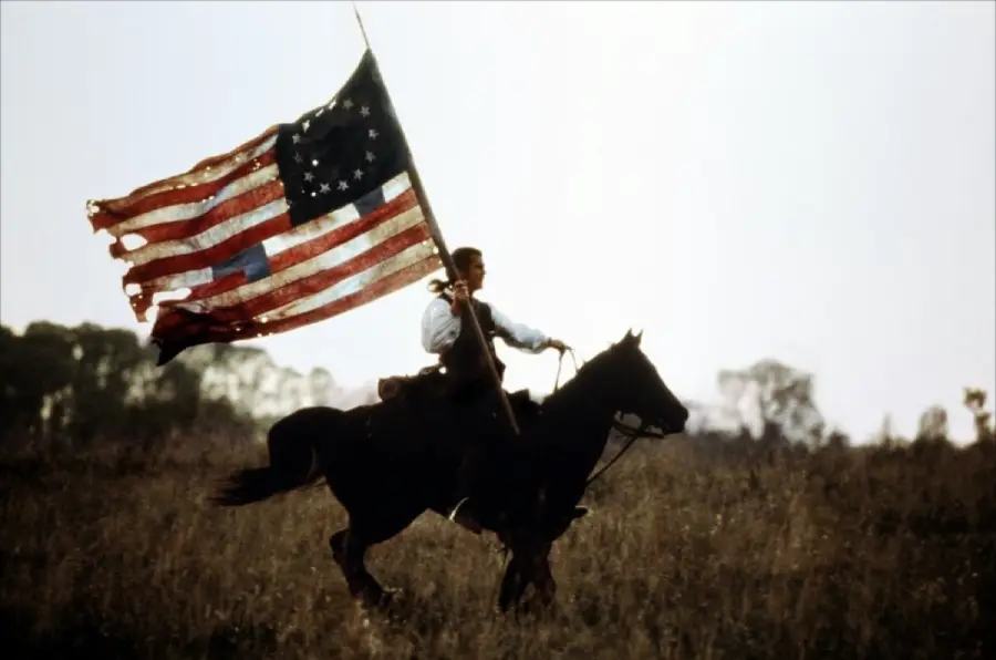 A person riding a horse through a grassy field, holding a large, tattered American flag with a circle of stars. The background is hazy with silhouettes of trees.