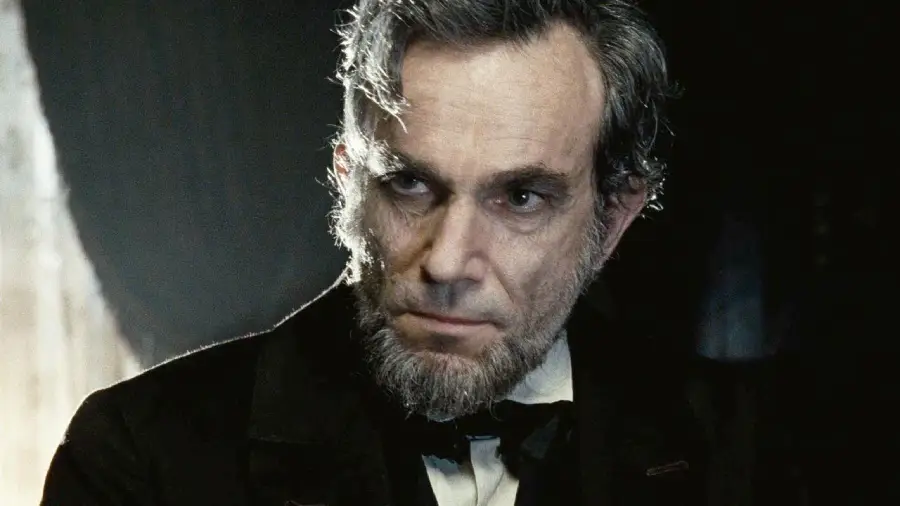 A man with gray hair, a beard, and a serious expression wears a black coat and bow tie, illuminated by soft lighting against a dark background.