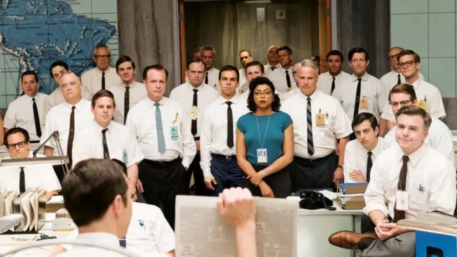A group of people in business attire, mostly men in white shirts and ties, stand closely together in an office with world maps on the walls, intently listening to a seated person holding papers.
