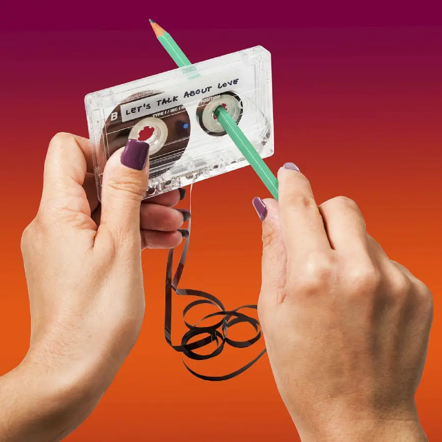 Two hands use a green pencil to manually rewind loose tape inside a clear cassette labeled "Let's Talk About Love" against a pink and orange gradient background.