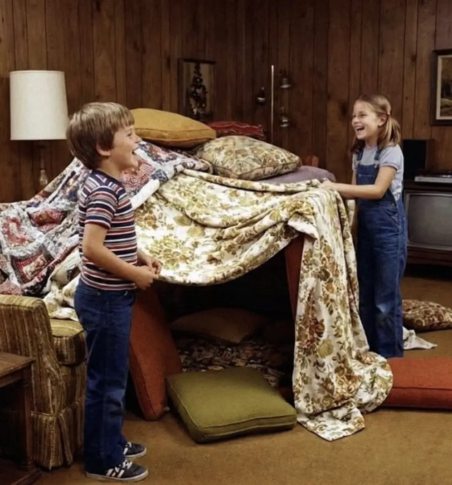 Two children smile and play in a living room, standing beside a large homemade blanket fort made of patterned blankets, cushions, and pillows. The room has wood-paneled walls and vintage furniture.