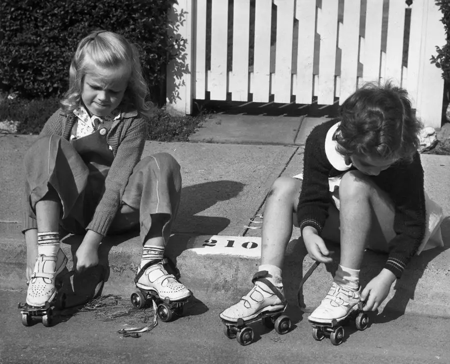 Two young girls sit on a curb in front of a white picket fence, putting on roller skates. One wears pants and a cardigan, the other a skirt and sweater. Both are focused on tying their skates.