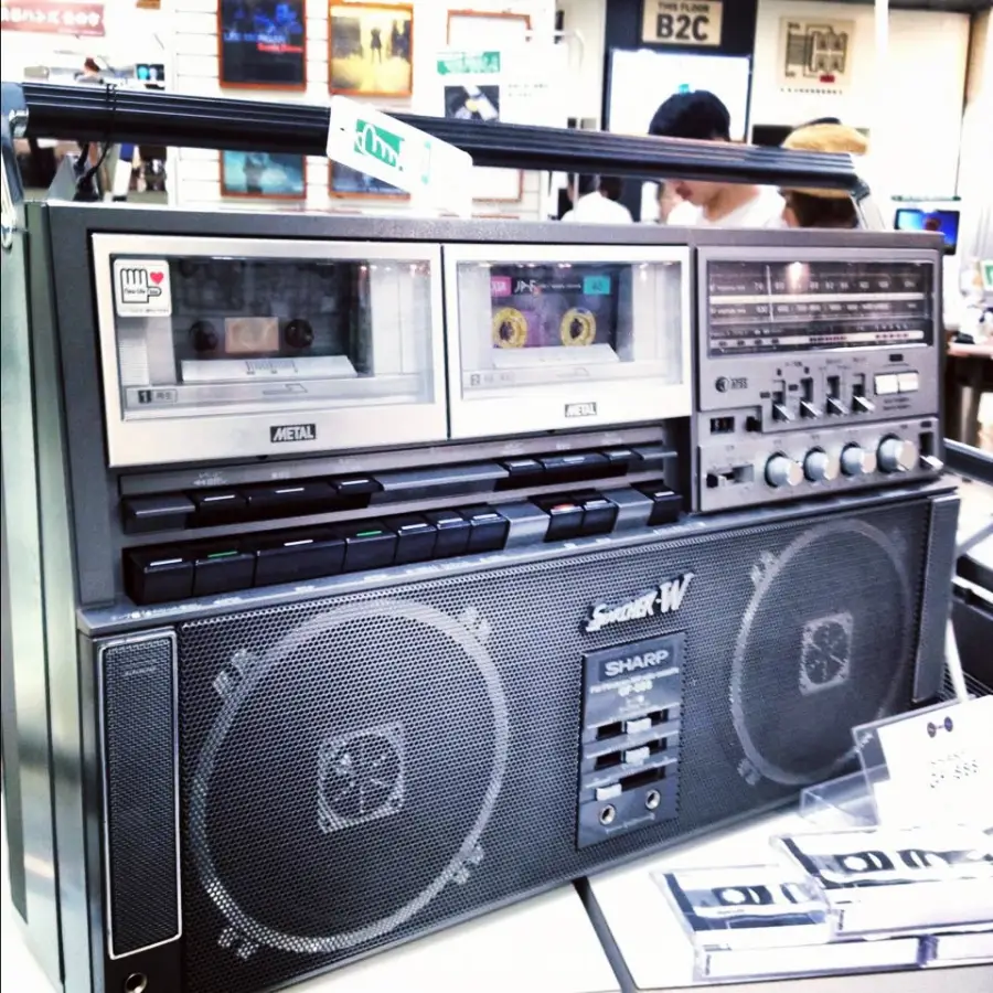 A vintage Sharp double cassette boombox with large speakers, various buttons, dials, and cassette tapes on display, sits on a counter in an electronics store. People and shelves are visible blurred in the background.