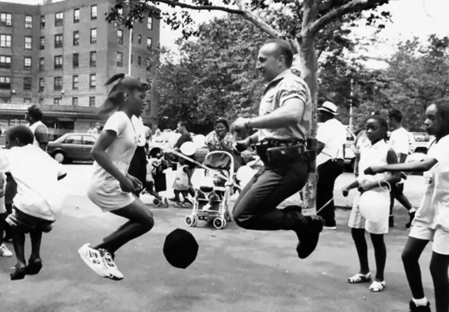 A police officer and a young girl are jumping rope together in a park, surrounded by smiling children and adults on a sunny day, with buildings and trees in the background.