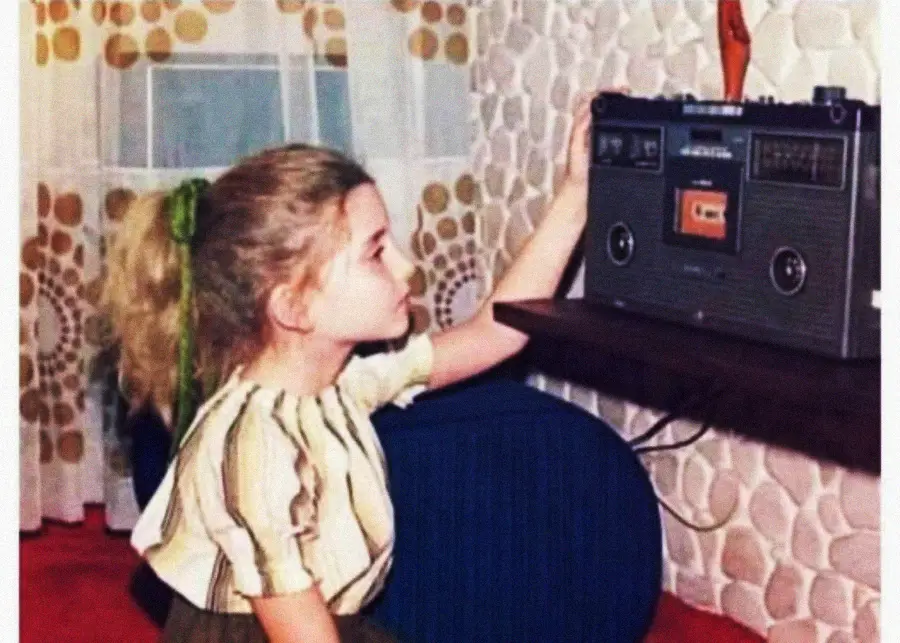 A young girl with blonde hair in a ponytail and a striped shirt is kneeling and reaching to operate a vintage cassette player in a room with patterned wallpaper and a window with sheer curtains.