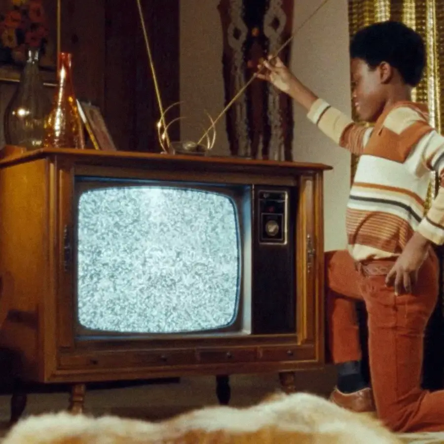 A young boy adjusts the antenna on a vintage wooden television displaying static in a retro-style living room decorated with patterned curtains and glass vases.