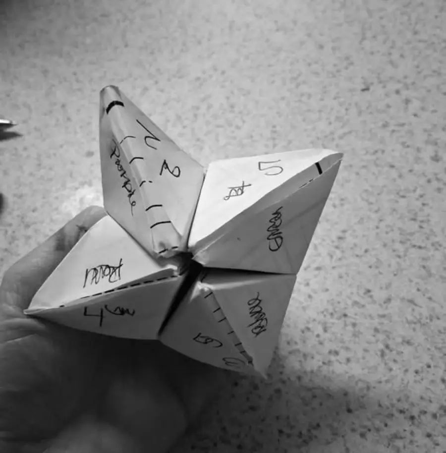 A hand holds a paper fortune teller with handwritten words and numbers on its flaps, resting on a speckled surface. The image is in black and white.