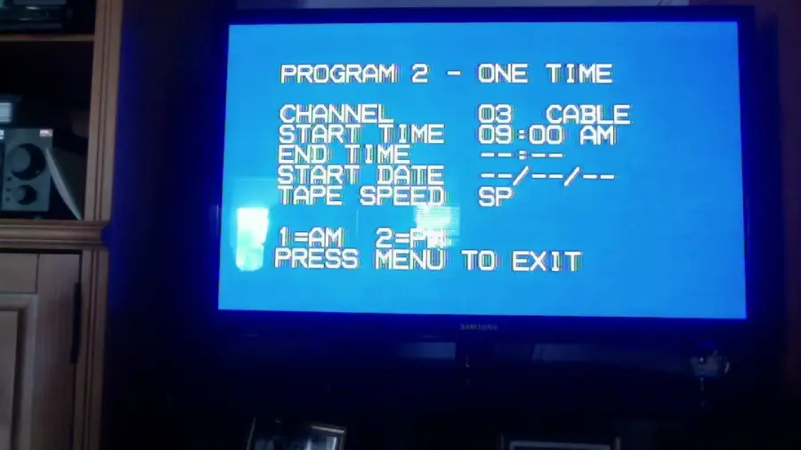 A television screen displays a blue menu for scheduling a recording: “PROGRAM 2 - ONE TIME, CHANNEL 03 CABLE, START TIME 09:00 AM, END TIME --:--, START DATE --/--/--, TAPE SPEED SP.” Prompts to press 1 or 2 to exit.