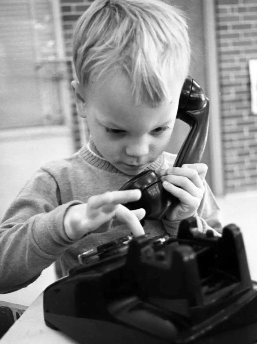 A young boy holds a rotary telephone receiver to his ear while using his finger to dial a number. He appears focused, with short blond hair, indoors near a brick wall.