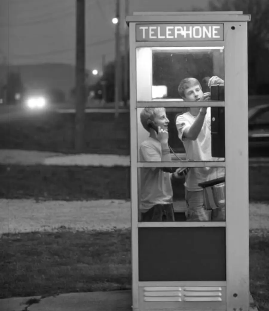 Two boys stand inside a glass phone booth at dusk; one boy is using the phone while the other is dialing. The booth is lit up and the word "TELEPHONE" is visible at the top. Car headlights shine in the background.