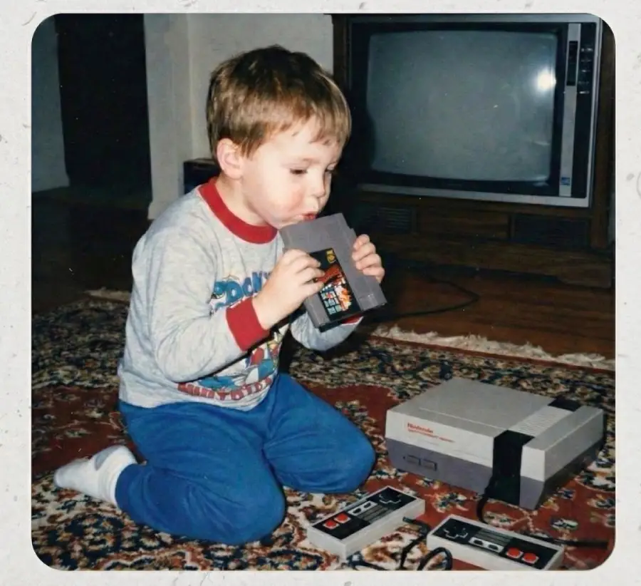 A young boy kneels on a carpeted floor, blowing into a Nintendo game cartridge beside an NES console and TV, with two controllers in front of him.