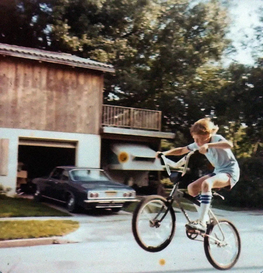 A young person in shorts and knee-high socks rides a BMX bike, popping a wheelie on a driveway in front of a two-story wooden garage with a car parked inside. Trees and greenery are in the background.