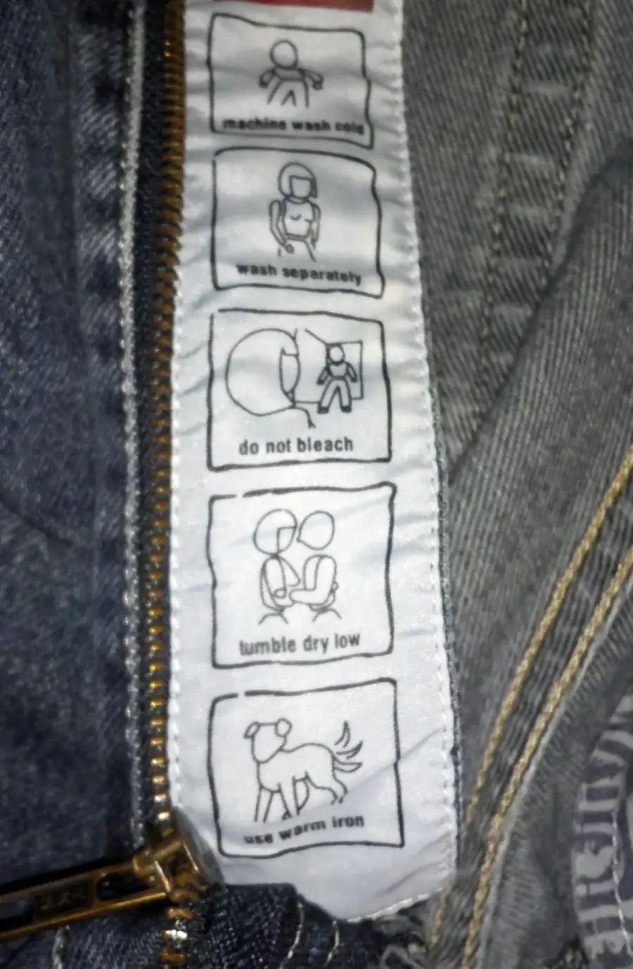 A clothing care label on denim jeans with five icons and instructions: machine wash cold, wash separately, do not bleach, tumble dry low, and use warm iron. The zipper and fabric texture are visible.