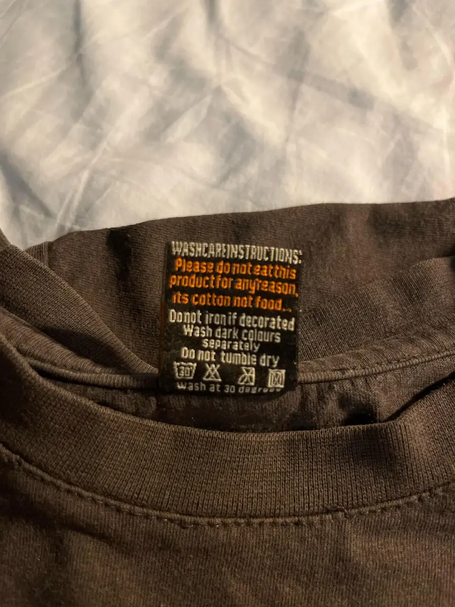 A close-up of a clothing tag on a dark t-shirt with humorous wash instructions, warning not to eat the product as it is cotton, not food, and displaying typical washing and drying symbols.