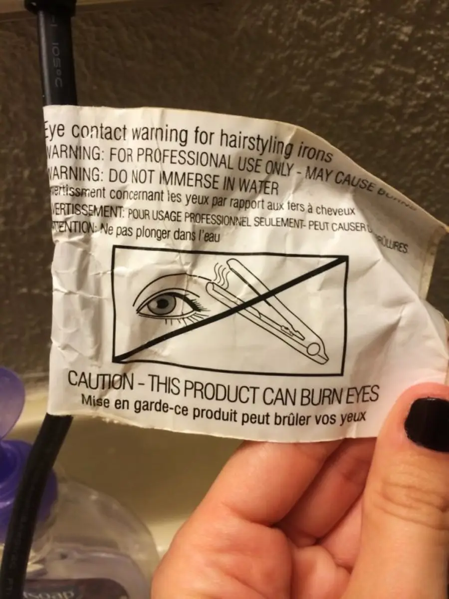 A hand holds a crumpled warning label with an illustration of an eye and a hair styling iron, crossed out. The label warns that the product can burn eyes and advises not to immerse it in water.
