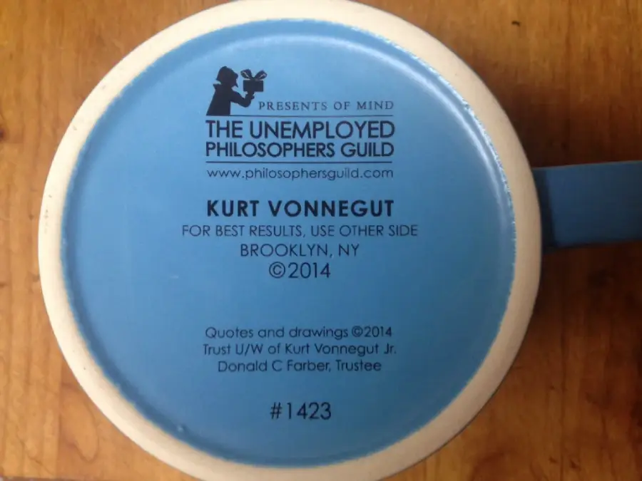 The bottom of a blue mug features The Unemployed Philosophers Guild logo, text referencing Kurt Vonnegut, a copyright notice from 2014, and the number 1423, all printed in black.