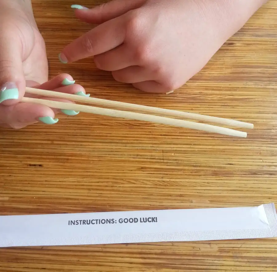 A person with mint green nail polish holds wooden chopsticks above a wooden table. Below, an empty chopsticks wrapper reads, "INSTRUCTIONS: GOOD LUCK!