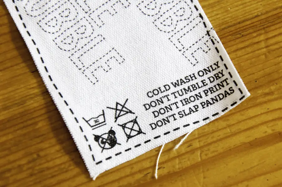 A white fabric tag on wood with printed care instructions: "COLD WASH ONLY, DON'T TUMBLE DRY, DON'T IRON PRINT, DON'T SLAP PANDAS," plus standard laundry symbols. Black dashed lines border the tag.