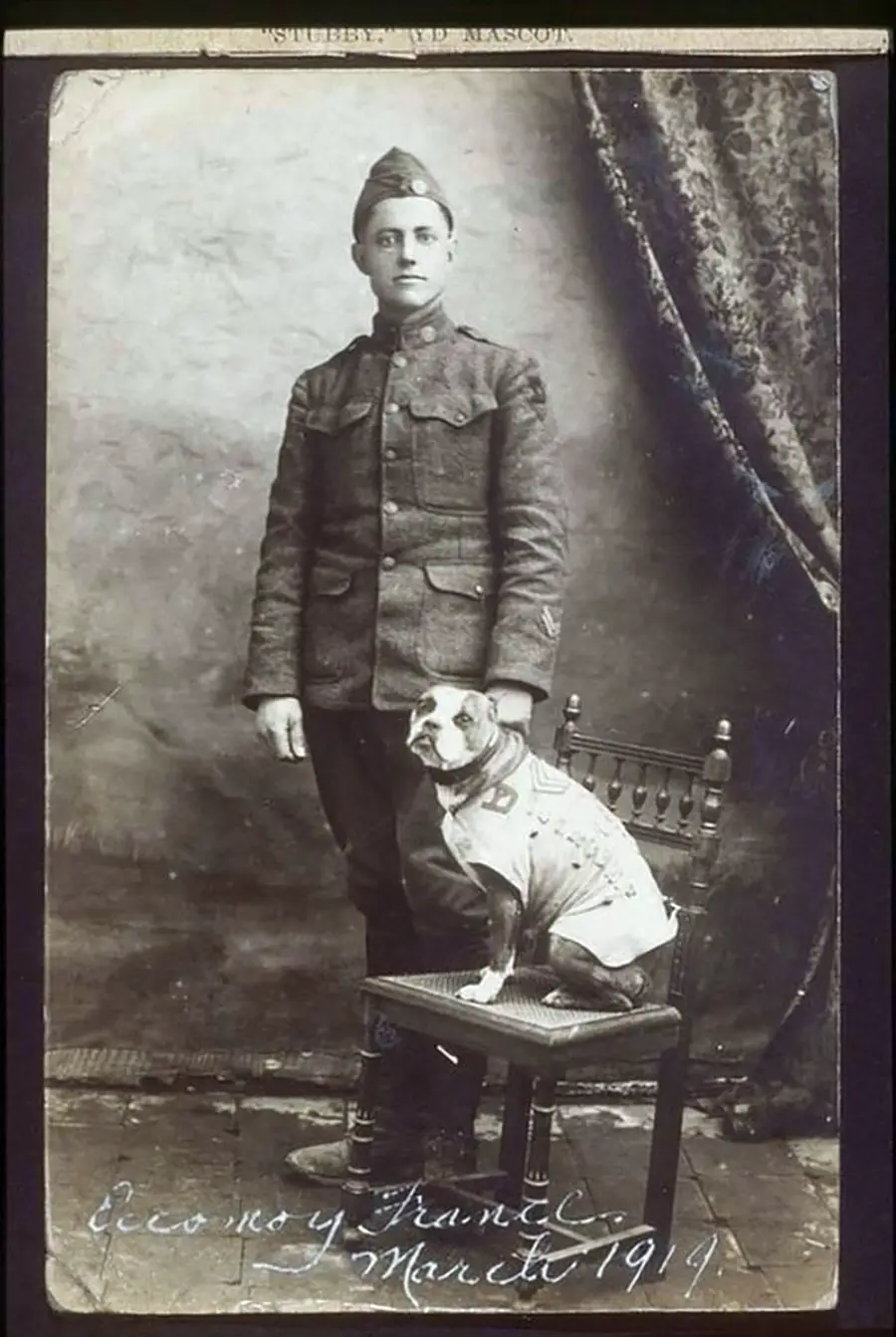 A black-and-white photo of a World War I soldier in uniform standing beside a wooden chair. A dog wearing a small coat sits on the chair. There is handwriting at the bottom, dated March 1919.