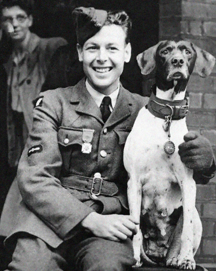 A smiling young man in a military uniform sits next to a dog with a collar and tag. The man has one arm around the dog, and another person stands blurred in the background.