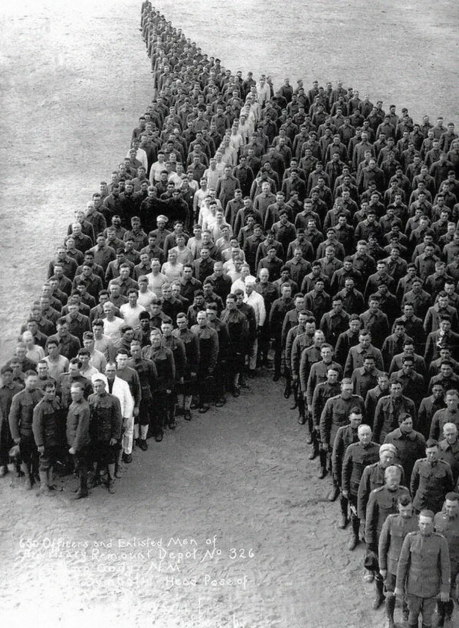 A large group of soldiers in uniform forms the shape of a giant human head facing left. The formation is viewed from above, showing hundreds of individuals standing in tightly organized rows.