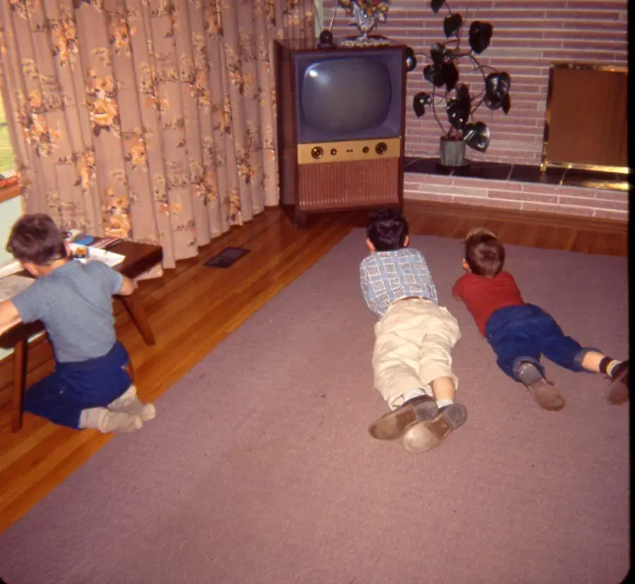 Three children are in a living room with wood floors; two boys lie on a rug watching a vintage TV, while another boy sits at a small table drawing. The room has floral curtains and a potted plant near the TV.