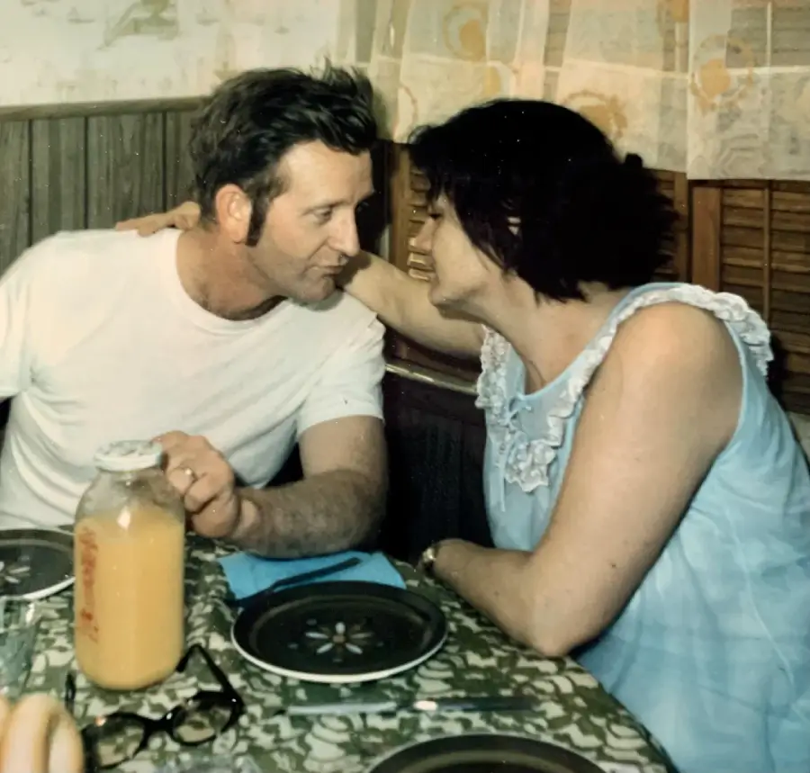 A man and woman sit closely at a table set for a meal, leaning in as if sharing an intimate conversation. The table has plates, a bottle of juice, and glasses. They appear relaxed and connected in a cozy, retro kitchen.