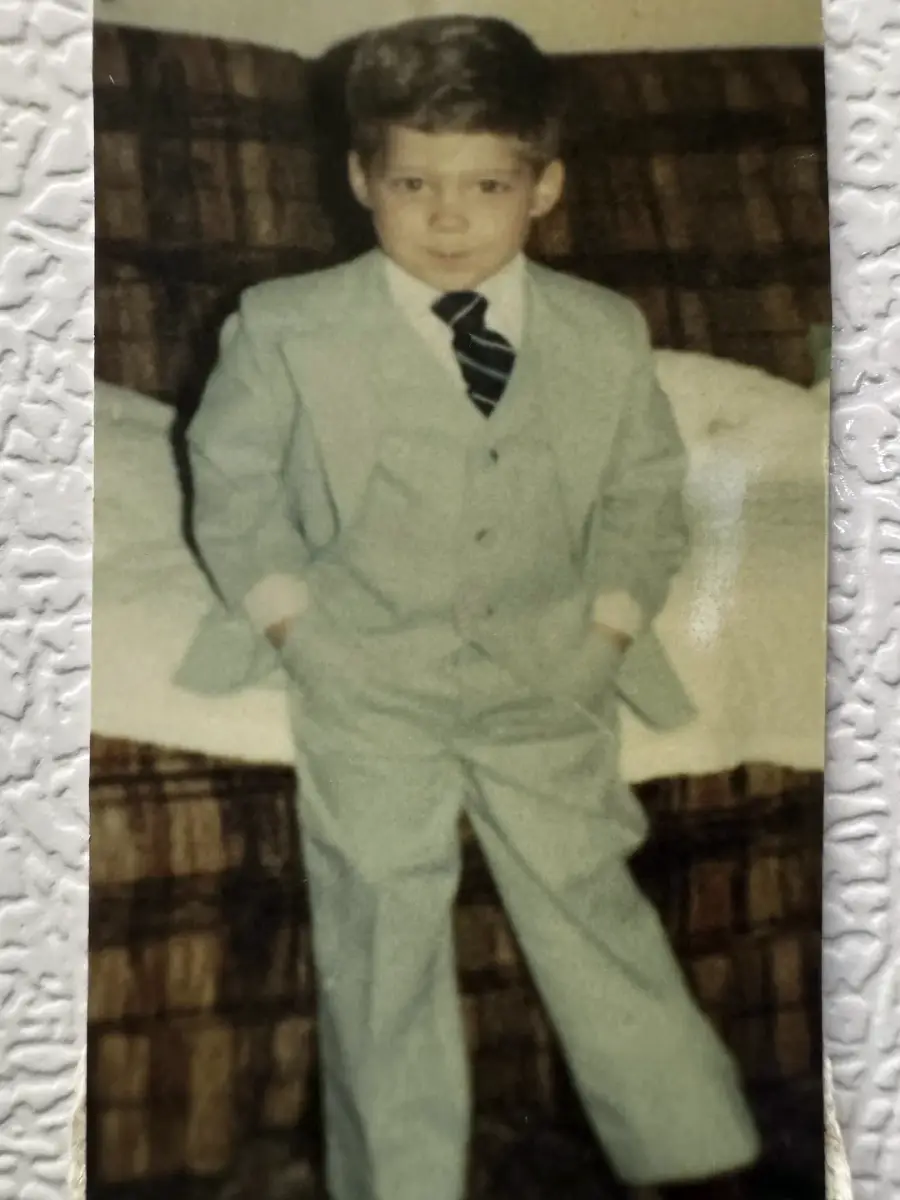 A young boy in a light gray suit and striped tie stands with his hands in his pockets in front of a brown plaid couch, looking at the camera with a slight smile.
