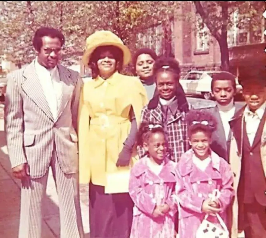 A family of eight poses outdoors on a sidewalk. The adults wear formal 1970s clothing, including a man in a striped suit and a woman in a yellow dress and hat. The children are dressed in coats, with two young girls in matching pink outfits.